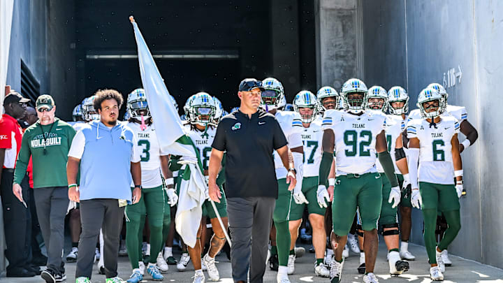 Tulane Green Wave football coach Jon Sumrall in black hat black shirt walks with team in white helmets white jerseys