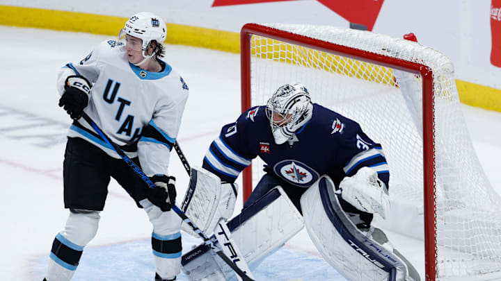 Nov 5, 2024; Winnipeg, Manitoba, CAN;  Winnipeg Jets goalie Connor Hellebuyck (37) and Utah Hockey Club forward Logan Cooley (92) look for the puck during the third period at Canada Life Centre. Mandatory Credit: Terrence Lee-Imagn Images