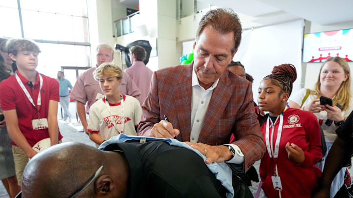 Aug. 5, 2025; Tuscaloosa, AL, USA; Nick Saban uses Cedric Burns’ back to help him sign autographs during the Nick’s Kids Foundation Luncheon at Bryant-Denny Stadium. Burns said they have been doing things this way for years when there is nothing else to sign on.