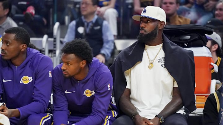 Oct 5, 2025; San Francisco, California, USA;   Los Angeles Lakers guard Bronny James (9) and forward LeBron James (23), wearing a Los Angeles Dodgers cap, watch the action against the Golden State Warriors in the first quarter at Chase Center. Mandatory Credit: David Gonzales-Imagn Images
