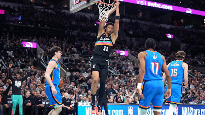 Feb 4, 2026; San Antonio, Texas, USA; San Antonio Spurs forward Carter Bryant (11) hangs on to the rim after dunking over Oklahoma City Thunder guard Brooks Barnhizer (23) during the first half at Frost Bank Center. Mandatory Credit: Scott Wachter-Imagn Images