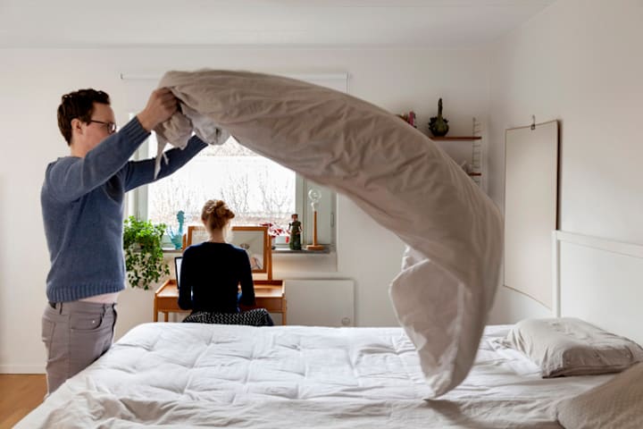 A man lifts a comforter over a bed to air it out while a woman sits at a computer desk in the same room