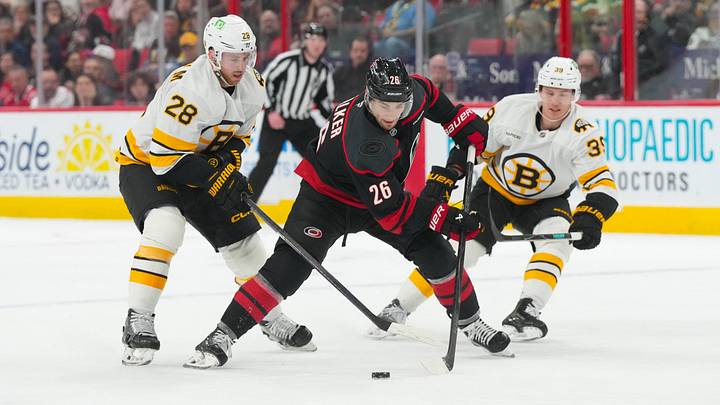 Apr 7, 2026; Raleigh, North Carolina, USA;  Carolina Hurricanes defenseman Sean Walker (26) skates with the puck between Boston Bruins center Elias Lindholm (28) and center Morgan Geekie (39) during the third period at Lenovo Center. Mandatory Credit: James Guillory-Imagn Images