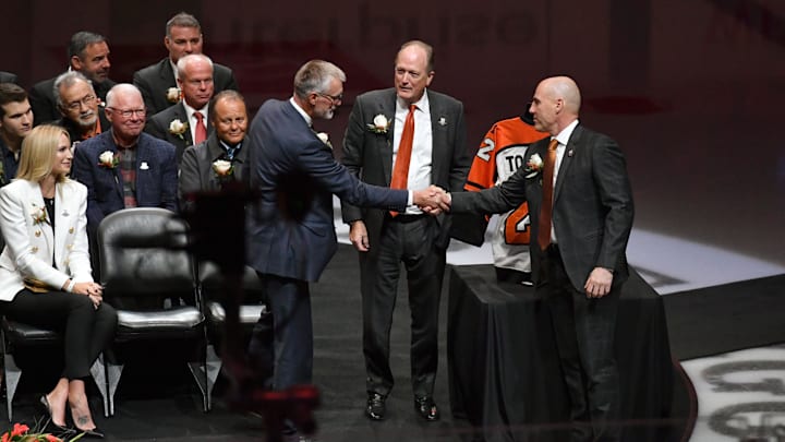 Nov 16, 2021; Philadelphia, Pennsylvania, USA; Former Philadelphia Flyers Paul Holmgren, left, shakes hands with Rick Tocchet after they were inducted to the Flyers Hall of Fame during a ceremony before game against the Calgary Flames at Wells Fargo Center. Dave Scott, Comcast Spectacor Chairman and CEO, and Governor of the Flyers stands between them.