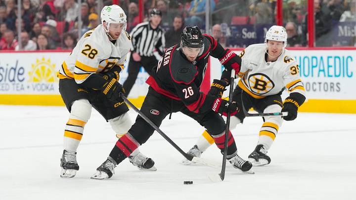 Apr 7, 2026; Raleigh, North Carolina, USA;  Carolina Hurricanes defenseman Sean Walker (26) skates with the puck between Boston Bruins center Elias Lindholm (28) and center Morgan Geekie (39) during the third period at Lenovo Center. Mandatory Credit: James Guillory-Imagn Images