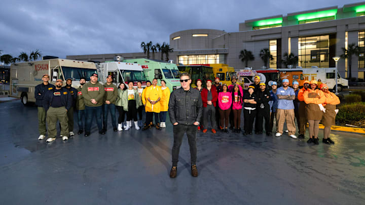 Nishaan, Fat Kid Food Co., Stop Drop N Roll, Rising Tiger, Good Fortune Company, Eat My Biscuits, Burger Walla, G’s Cheesesteaks and Cooking with Que team members in front of their trucks with host Tyler Florence, as seen on The Great Food Truck Race, Season 18.
