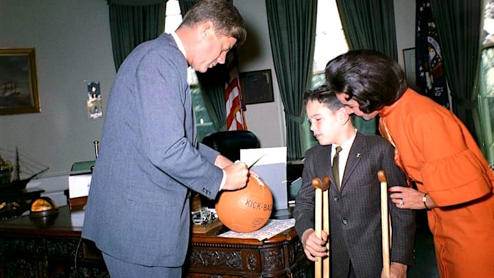 President John F. Kennedy signs a kick ball for Thomas A. Doyle III, the 1962 Easter Seal Child. Pauline Phillips (a.k.a. Dear Abby) is at right.