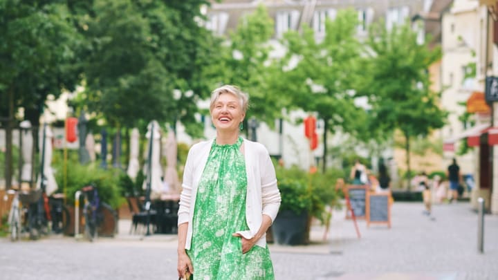 Older woman in green dress smiling on a city street with green trees