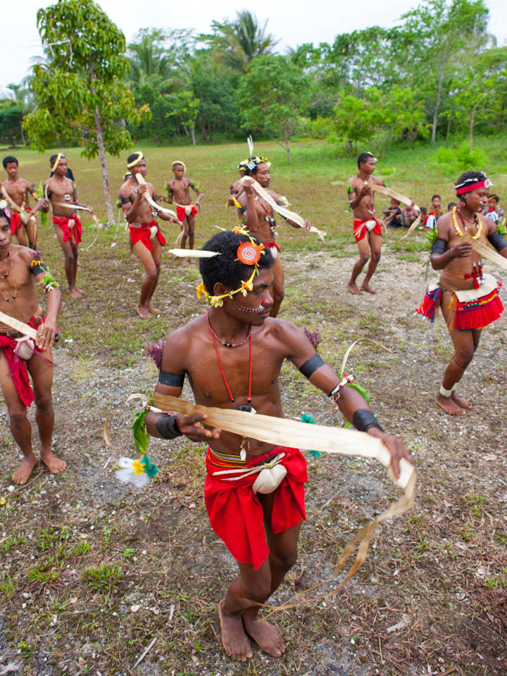 about ten Trobriand Island dancers dressed in red bottoms, black arm bands, jewelry, and headgear 