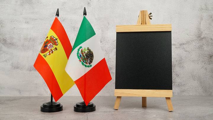 Spain and Mexico flags beside a blank chalkboard on a neutral background with copy space. Spain and Mexico flags beside a blank chalkboard on a neutral background with copy space.