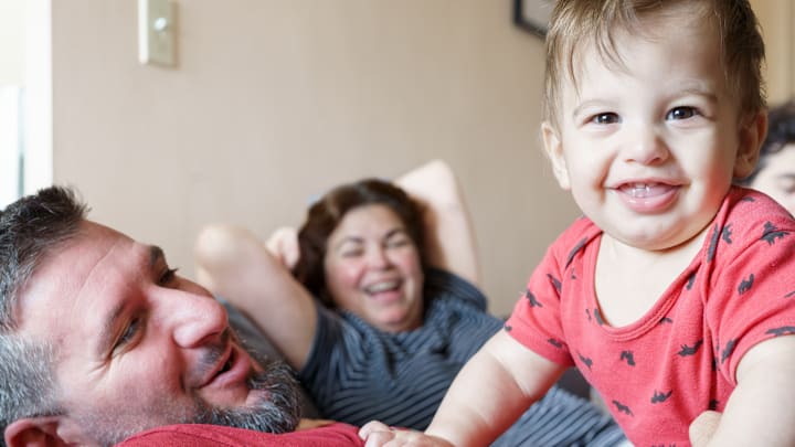 Smiling baby in red outfit leans over man as woman laughs in Smiling baby in red outfit leans over man as woman laughs in