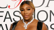 Serena Williams poses in front of a white and black backdrop in a black dress and floral diamond necklace.