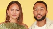 Chrissy Teigen and John Legend pose side-by-side in front of a yellow backdrop. Teigen wears a green pleated dress and Legend wears a white tee and cream-colored sweater.