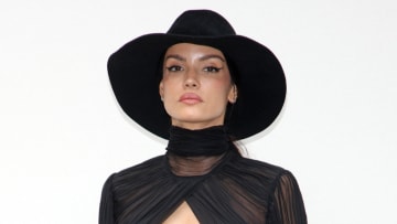 Sofia Resing poses in front of a white background in a high neck black sheer shirt and a wide-brimmed hat.