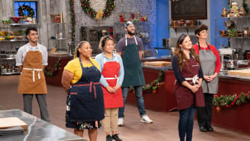 Bakers Loreno, LaShonda, Julianna, Jon, Megan and Eva learning about main heat bake, as seen on Holiday Baking Championship, Season 7. Photo provided by Food Network