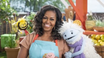 Michelle Obama, wearing a blue apron, stands in a rooftop garden posing for the camera and holding a pink, round mochi puppet. A bee puppet wearing a red tie hovers over her shoulder while a furry white and blue puppet with frozen waffle ears embraces her side.