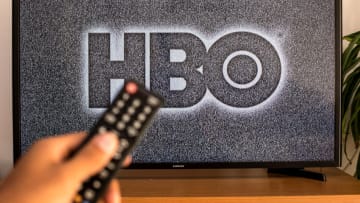 SPAIN - 2021/07/13: In this photo illustration a close-up of a hand holding a TV remote control seen displayed in front of the HBO logo on a tv in the background. (Photo Illustration by Thiago Prudencio/SOPA Images/LightRocket via Getty Images)