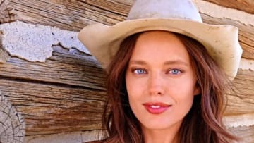 Emily DiDonato poses in front of a wooden cabin wearing a red swimsuit and a tan cowgirl hat.