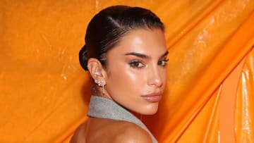 Jessica Aidi wears a slicked-back bun and a gray halter top and poses in front of an orange backdrop.