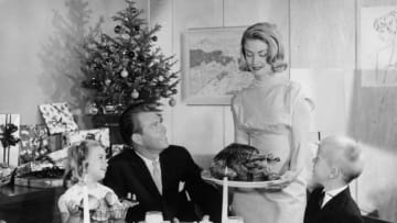 1965: An American family admire a turkey their mother is bringing to the table. A small Christmas tree with presents is in the background. (Photo by Keystone/Getty Images)