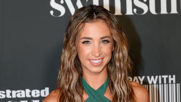 Katie Austin wears a green halter neck dress and her dirty blonde hair down in a beach wave, smiling at the camera in front of a black backdrop.