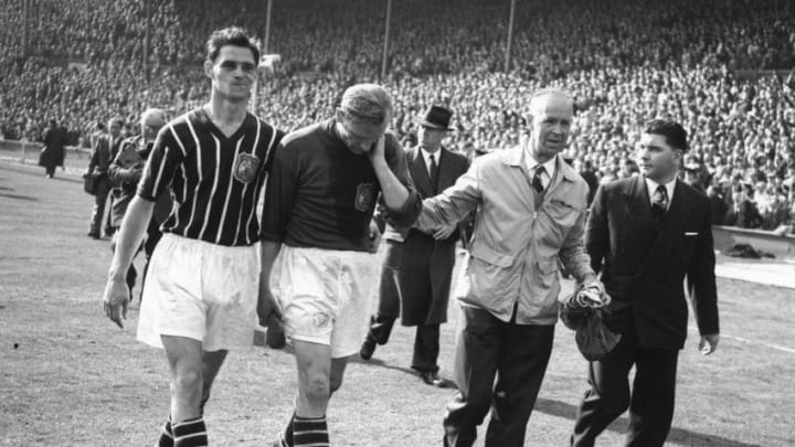 5th May 1956: FA Cup Final: Birmingham City v Manchester City. After helping Manchester City win the Cup 3-1 Bert Trautmann, injured during the match, is helped off the field rubbing his 'sore'' neck. It later transpired he had broken it. Trautmann was later named as player of the year. Mandatory Credit: Allsport Hulton/Archive
