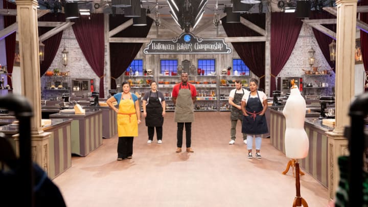 Bakers Michelle, Renee, Michael, Aaron and Sinai standing in kitchen with costumes in foreground, as seen on Halloween Baking Championship, Season 6., as seen on Halloween Baking Championship, Season 6. Photo provided by Food Network