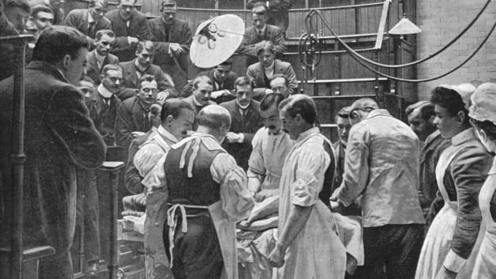 A team of surgeons work on a patient in the operating theater of London's Charing Cross Hospital while men in the gallery observe the procedure.