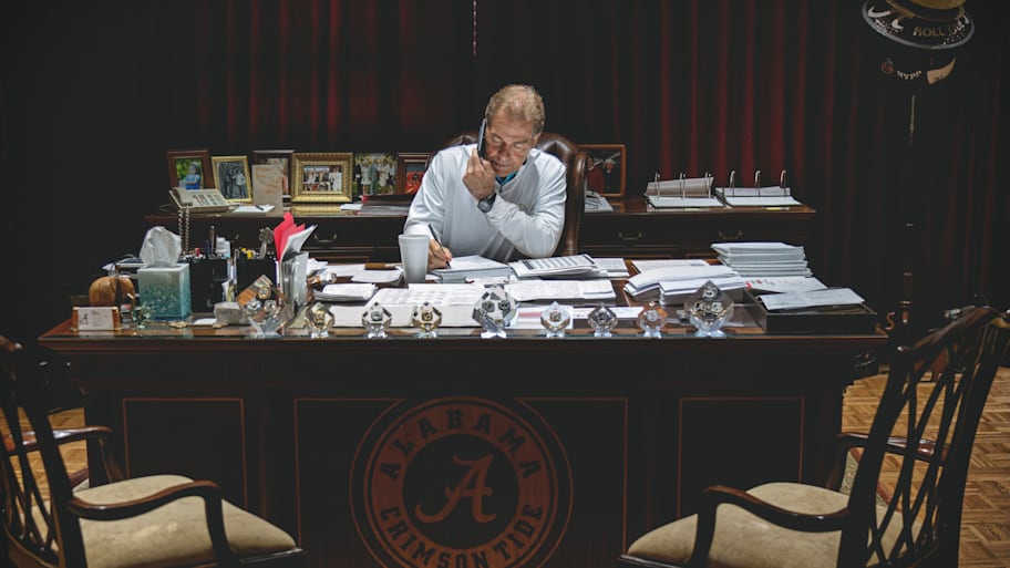 Nick Saban in his Alabama football office speaking on the phone while writing on a paper.