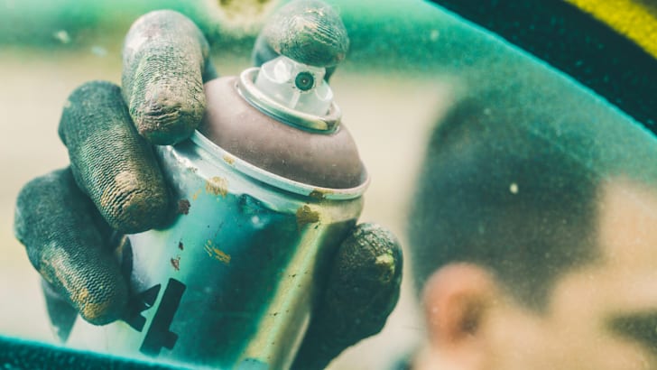 A gloved hand holding a spray paint can with a finger on the trigger.