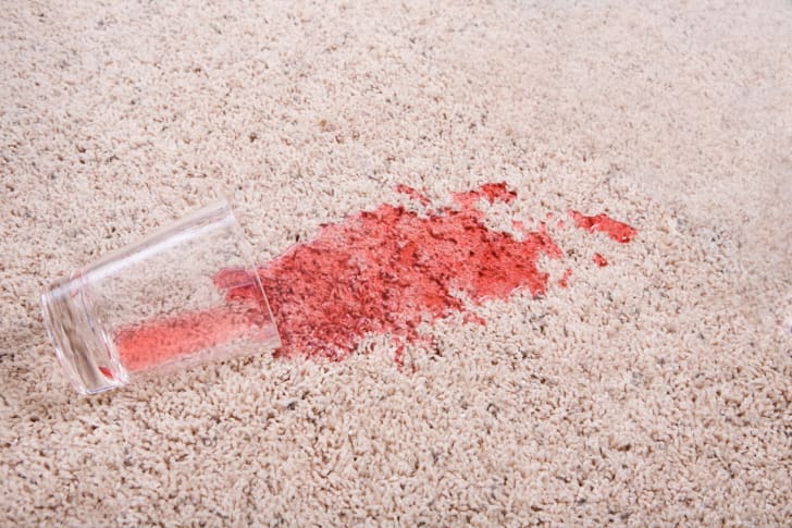 A clear glass tipped sideways on an off-white colored carpet, spilling red juice out onto the rug.