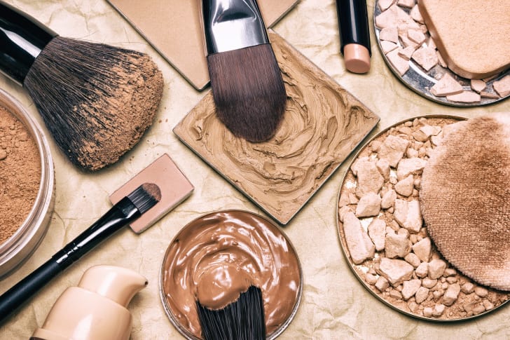 Various circular and square pans containing liquid and powder makeup, with brushes dipping into some of them.