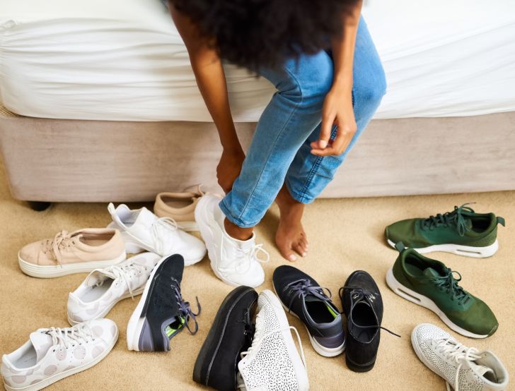 Woman sitting on bed trying on shoes from pile