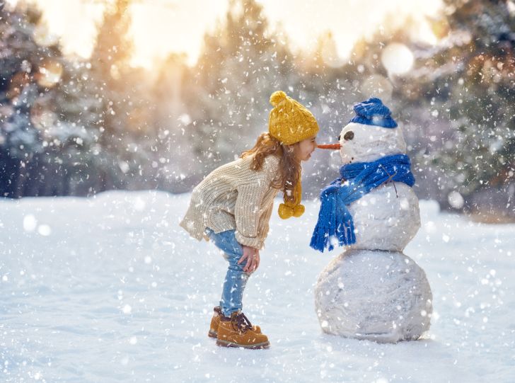 A little girl rubbing her nose on the carrot nose of a snowman while snow falls.