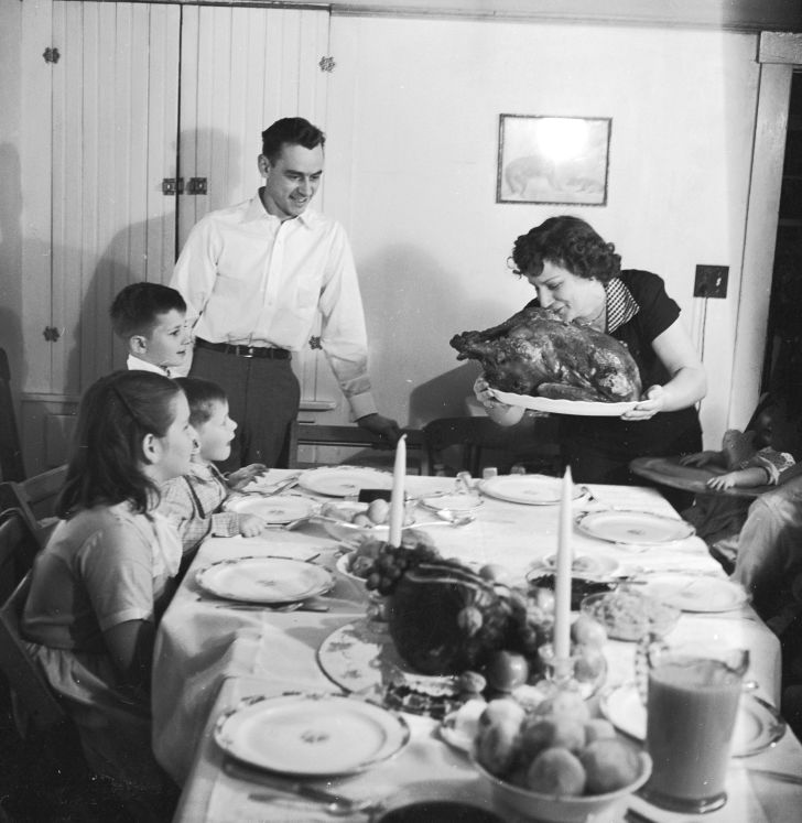 A black and white photo of a family gathering around the table as the mother brings in a turkey.