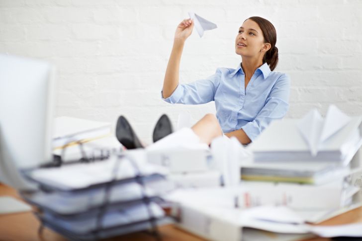 A young woman at a desk surrounded by paperwork, playing with a paper airplane
