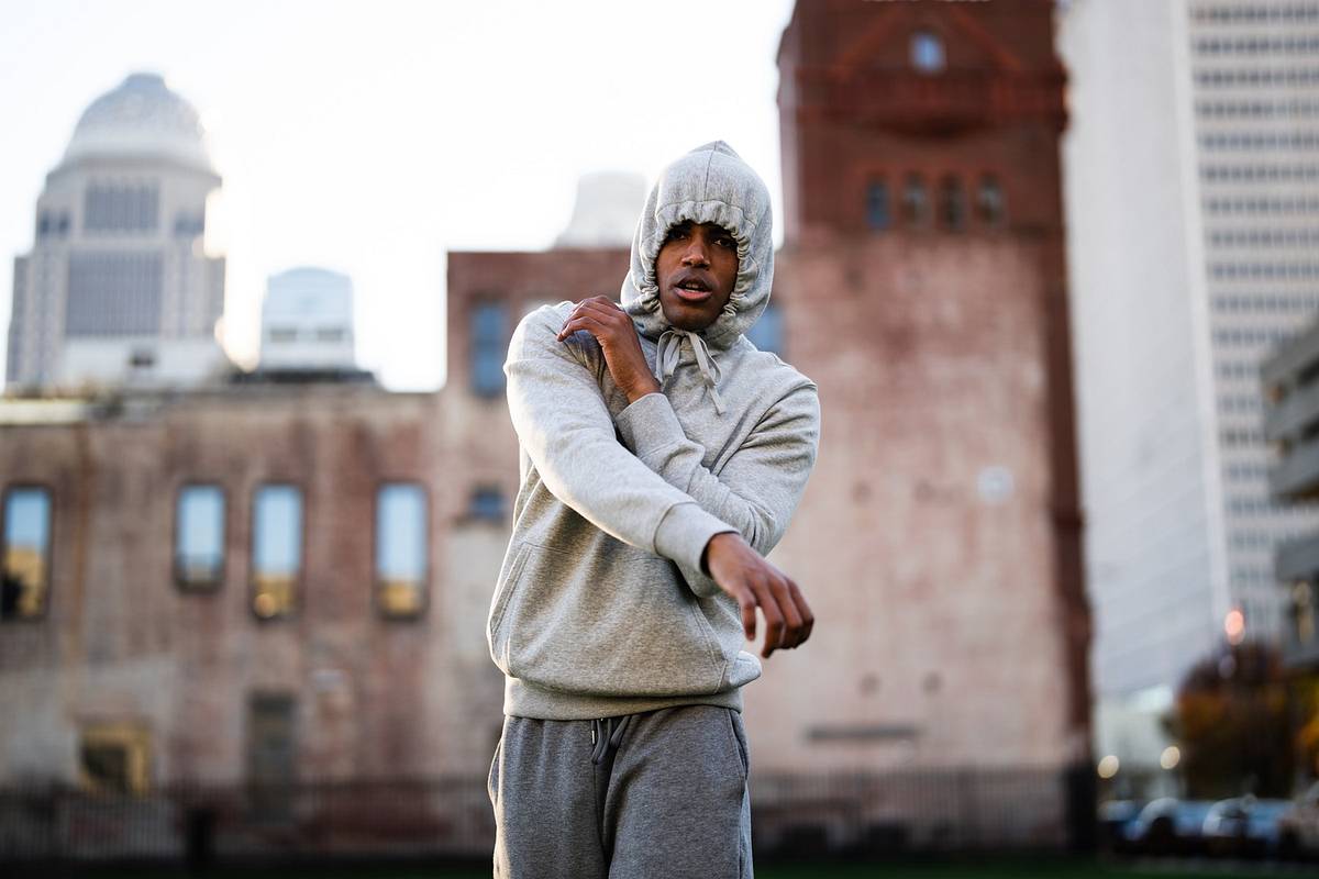 An actor in a gray hooded sweatsuit stretches his arm on a Louisville street, with brick buildings and downtown towers blurred behind him.