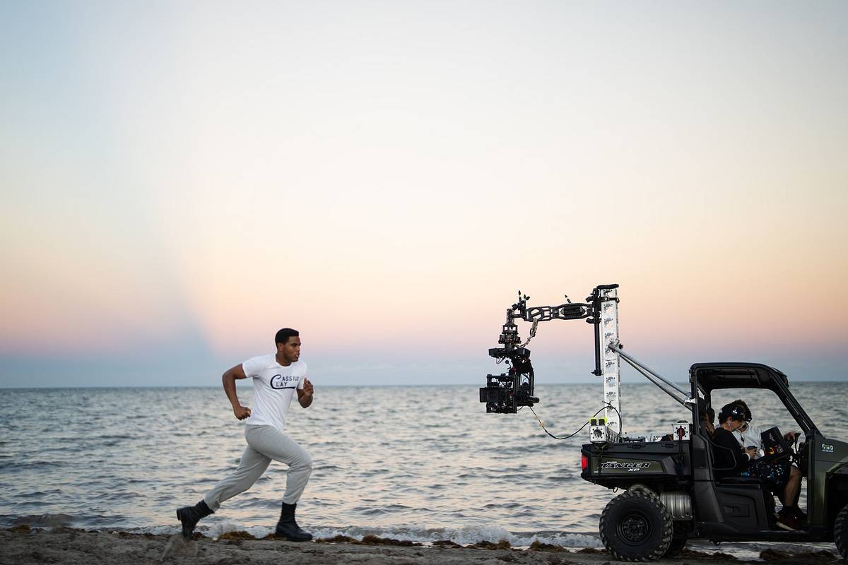 An actor in a white T-shirt runs along the shoreline at dusk while a camera crane mounted on a utility vehicle films alongside him.