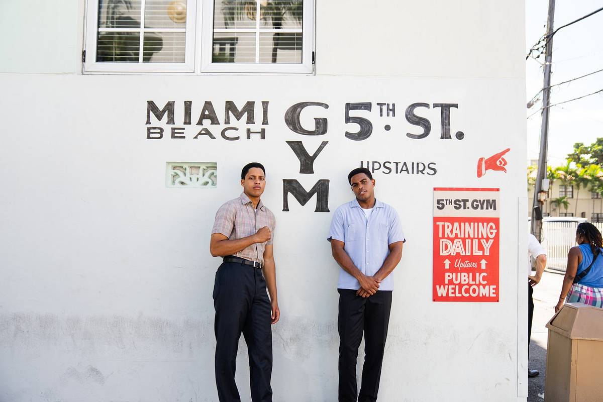 Two actors in 1960s clothing stand against a white wall painted with "Miami Beach 5th St. Gym" and a red "Training Daily, Public Welcome" sign.