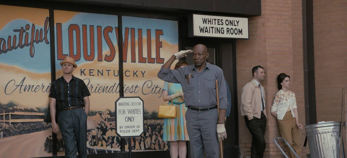A film still of a 1960s Louisville street, where costumed extras stand outside a "Whites Only Waiting Room" beside the "Beautiful Louisville" mural as one man salutes.