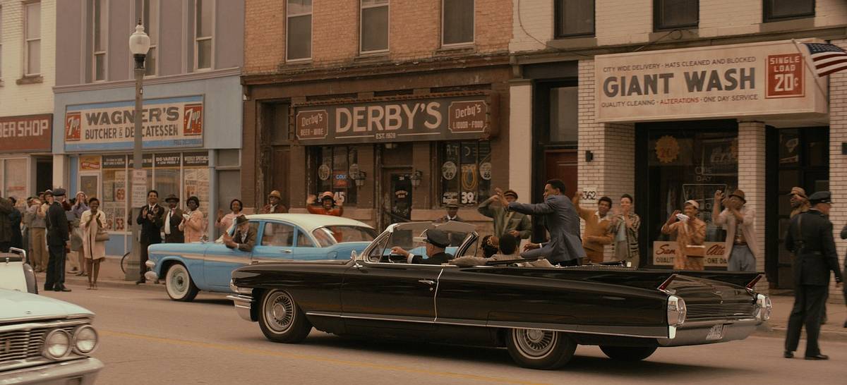 A film still of a period Louisville street, where crowds line the sidewalk as a black 1960s convertible drives past storefronts including Derby's and Giant Wash.
