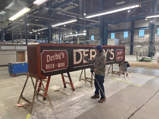 A crew member paints a large vintage-style "Derby's Beer · Wine" sign set up on sawhorses in a warehouse workshop.