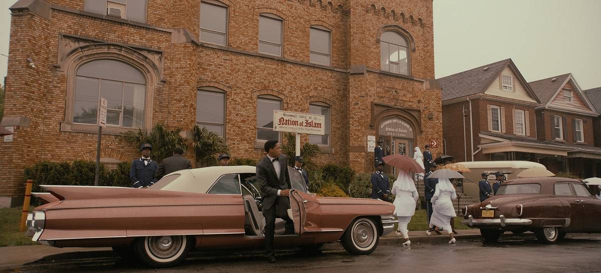 A film still of a rainy street outside a brick Nation of Islam building, where a man in a suit steps out of a pink 1960s Cadillac as women in white and uniformed men look on.