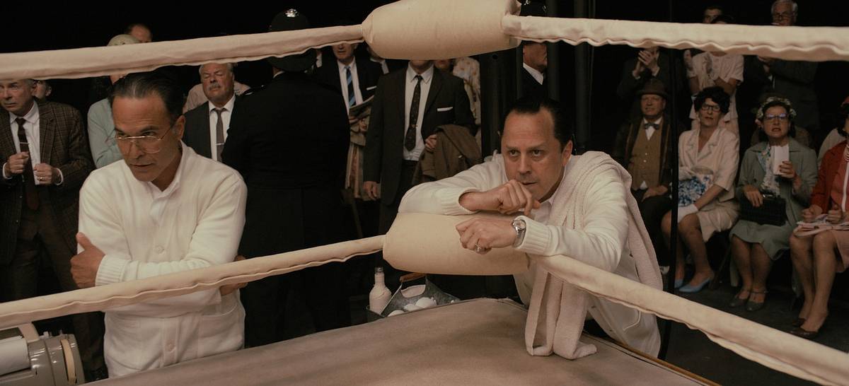 A film still of two cornermen in white jackets leaning on the ring ropes, with a crowd in 1960s dress behind them.