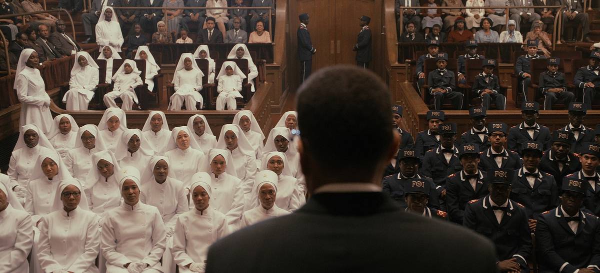 A film still of a man in a dark suit, seen from behind, facing rows of women in white and uniformed men in "FOI" caps.