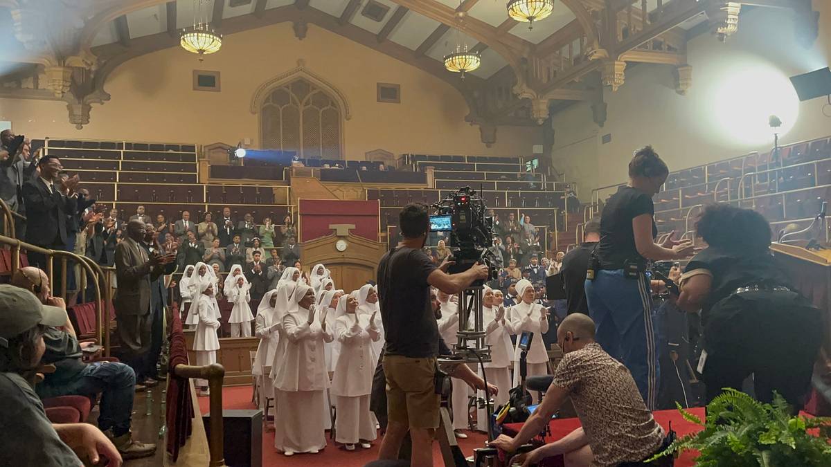 Behind the scenes in an ornate hall, a camera crew films a congregation of women in white robes and head coverings while others stand and applaud.