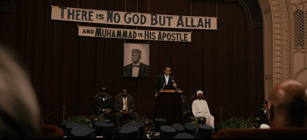 A film still of a speaker at a podium beneath banners reading "There Is No God but Allah and Muhammad Is His Apostle," with a portrait hanging behind him.