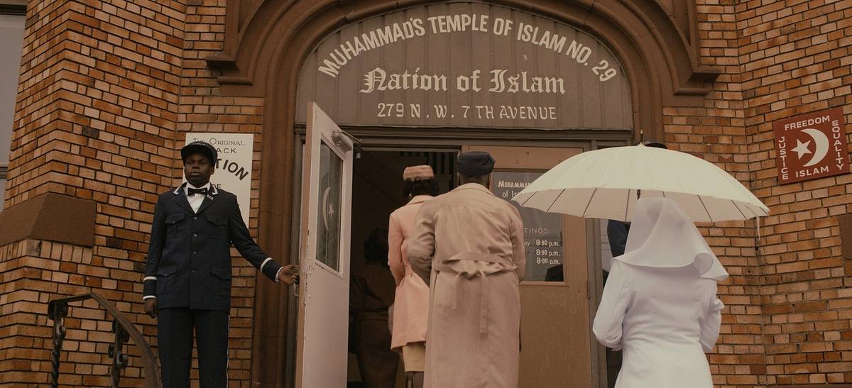 A film still of people entering "Muhammad's Temple of Islam No. 29, Nation of Islam," as a uniformed man holds the door and a woman in white carries a parasol.