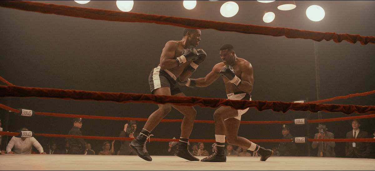 A film still of two boxers trading punches in a ring with red ropes beneath bright overhead lights.