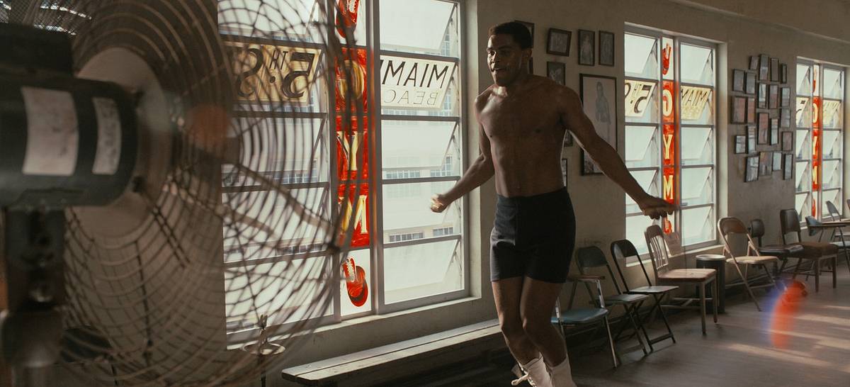 A film still of a shirtless boxer skipping rope in a sunlit gym, with a fan in the foreground and "Miami Beach 5th St. Gym" lettering on the windows.